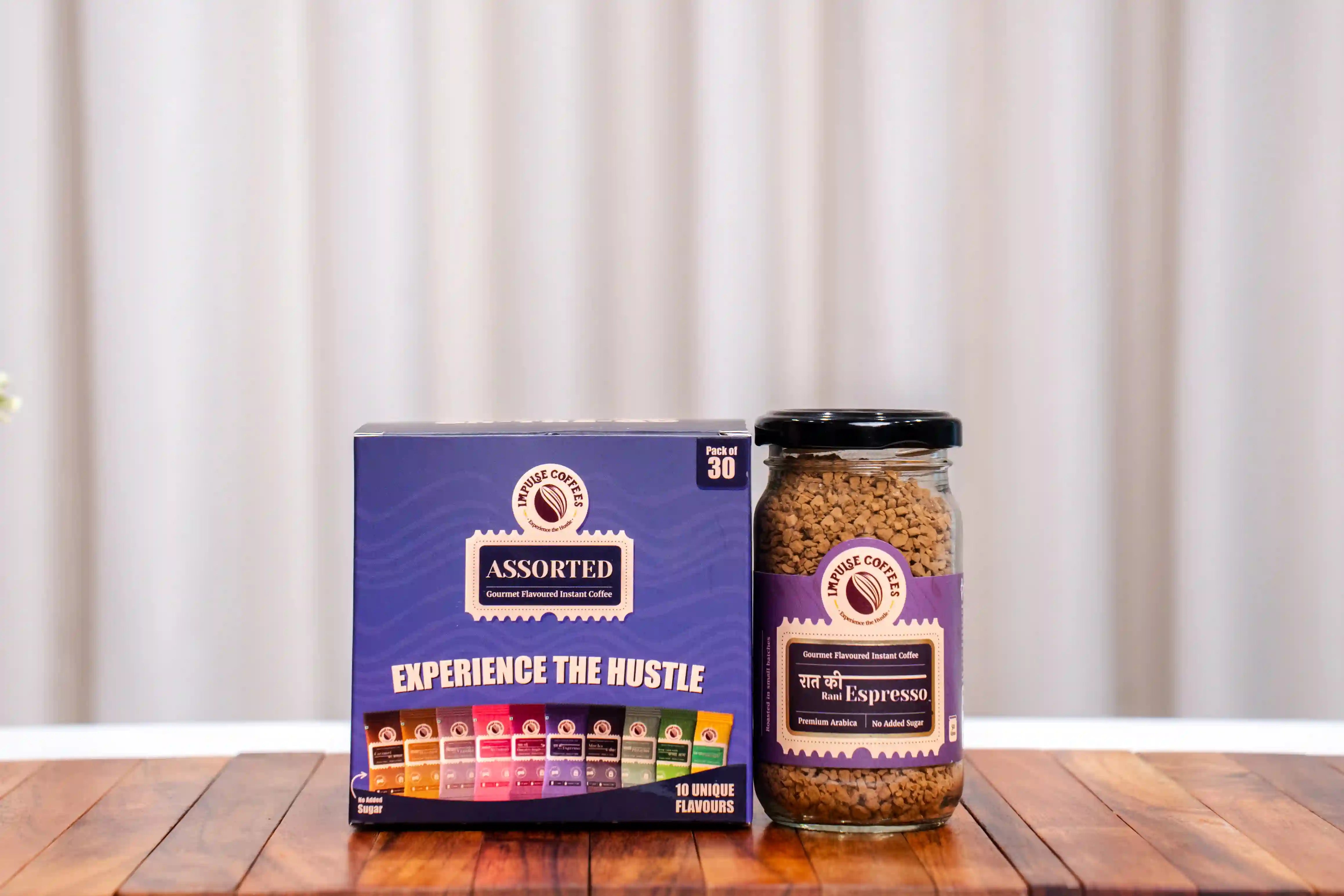 Assorted coffee pack of 30 and an espresso jar of 50gms on a wooden surface with a white curtain background